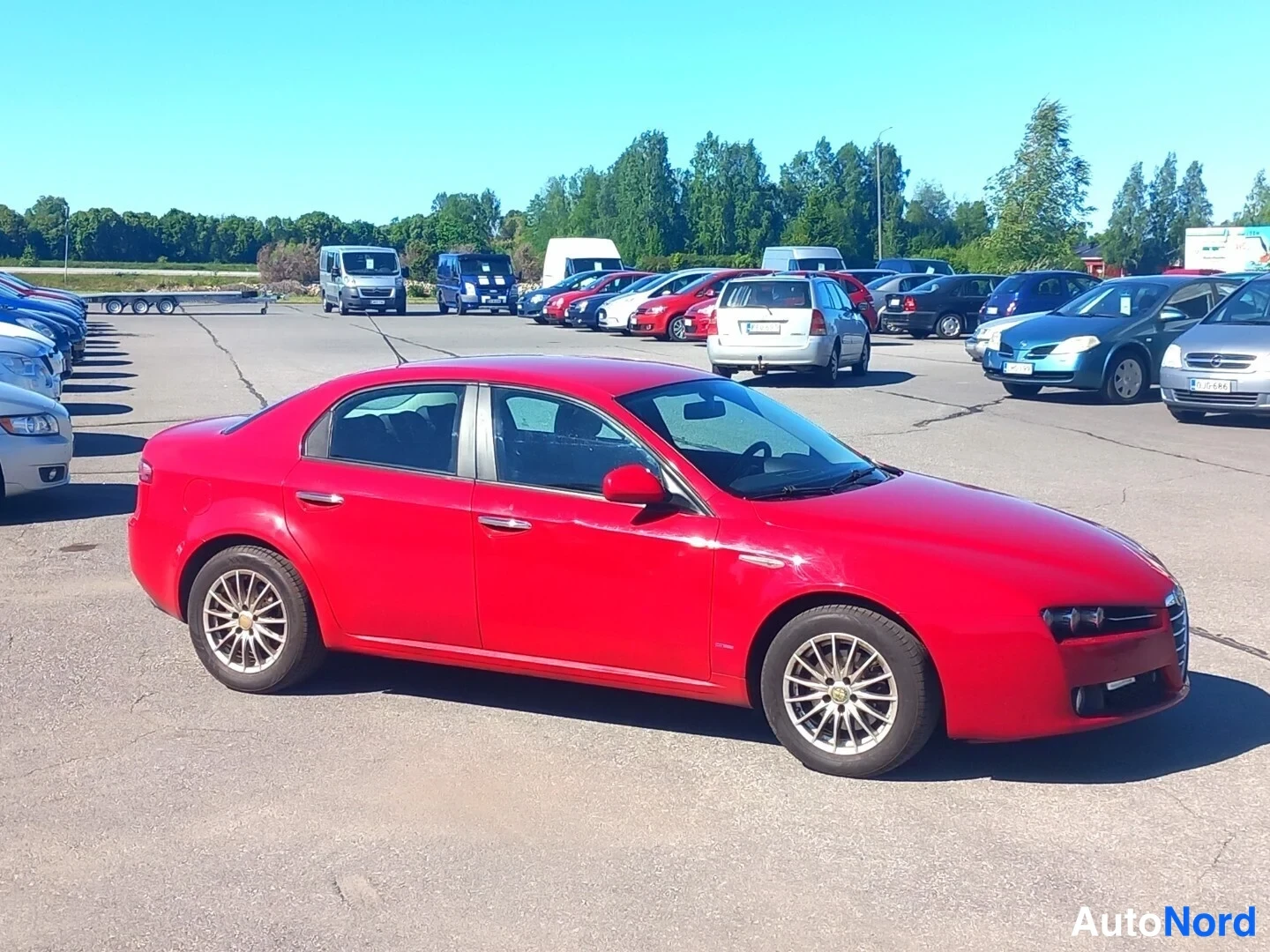 Etuviisto, nokka oikealle: Punainen Alfa Romeo 159 -sedan kuvattu kolmeneljänneltä edestä auton ollessa parkkipaikalla muissa autoissa.