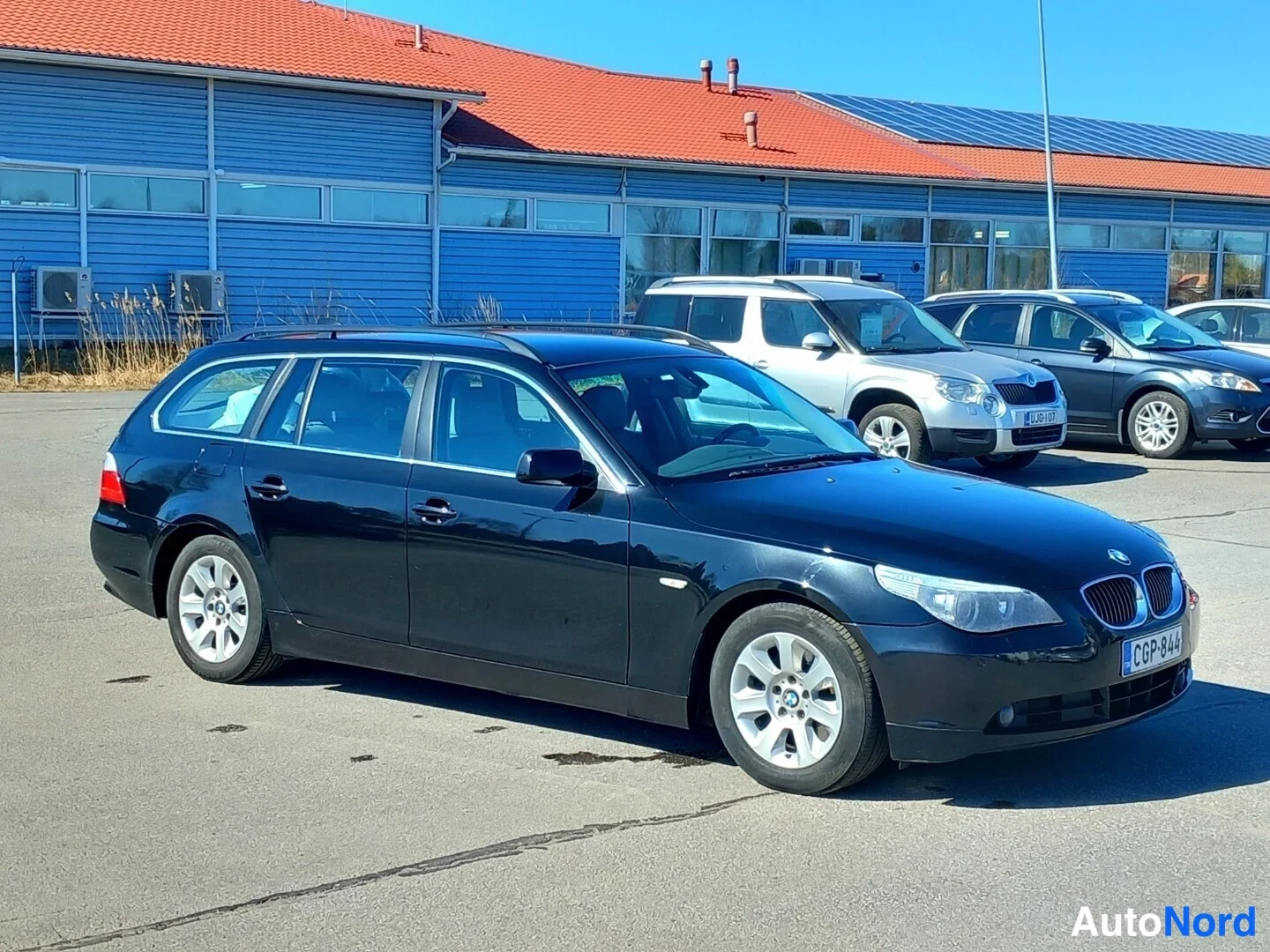 Etuviisto, nokka oikealle: Musta BMW-farmariauto kulmassa kuvattuna edessä ja oikealta sivulta, edessä BMW-merkitty etusäleikkö ja valkoinen rekisterikilpi.