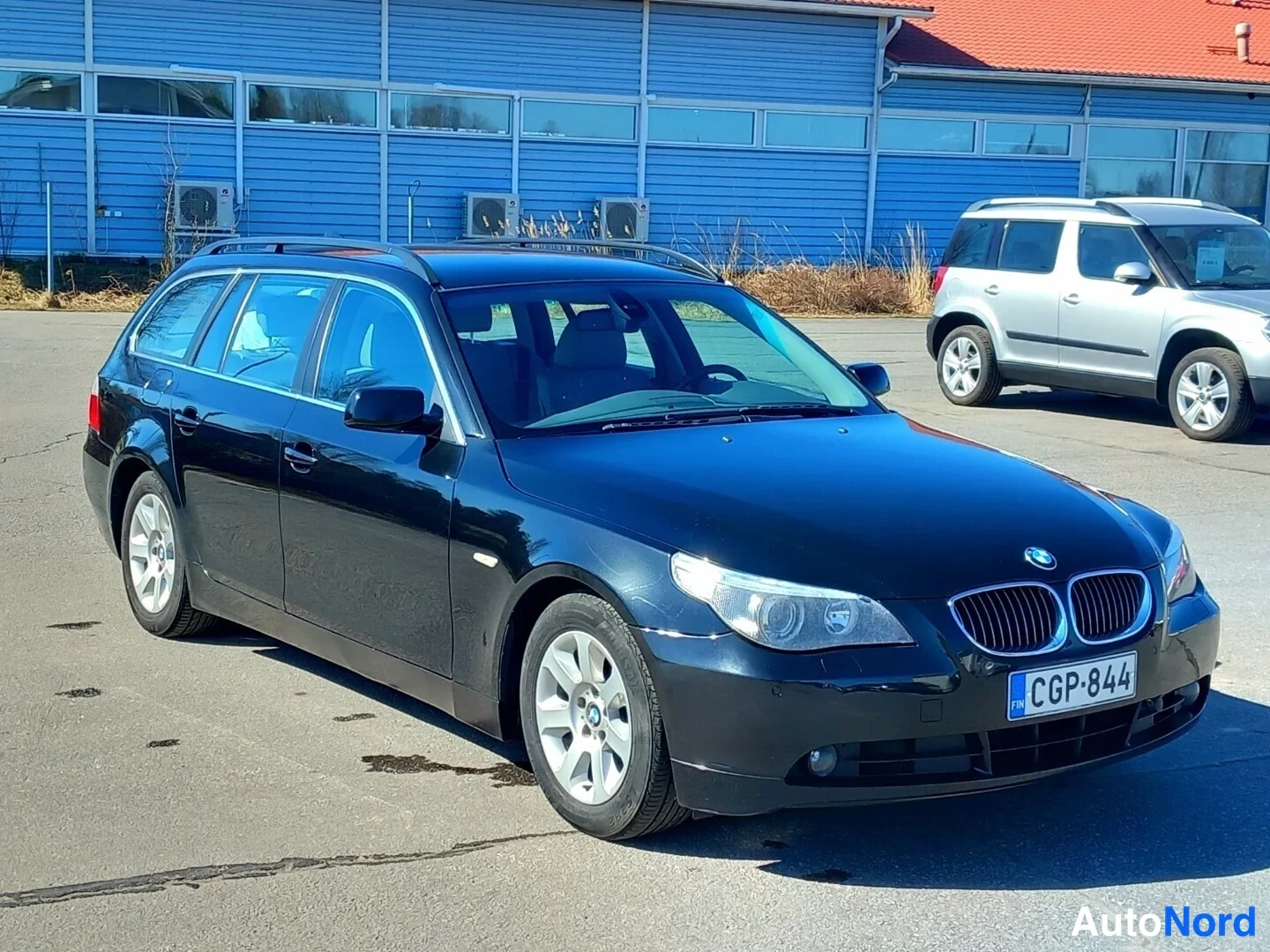 Etuviisto, nokka oikealle: Musta BMW-farmariauto kulmassa kuvattuna edessä ja oikealta sivulta, edessä BMW-merkitty etusäleikkö ja rekisterikilpi.