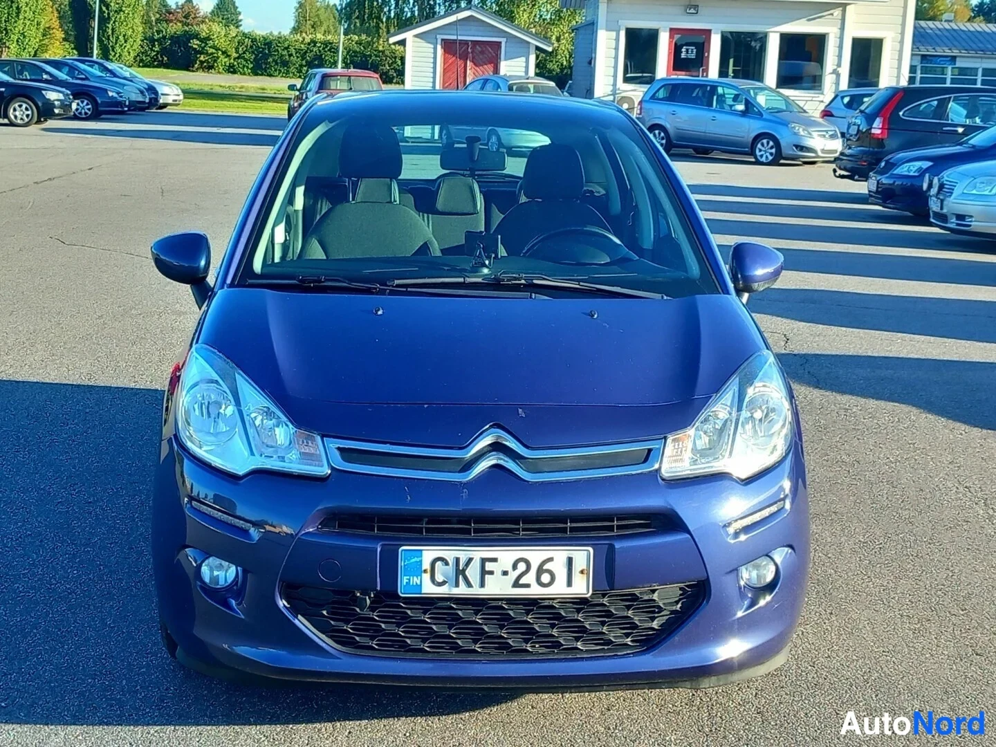 Edestä: Sinisen Citroën C3 -auton etunäkymä, rekisterikilpi, parkkialueella muiden autojen taustalla.