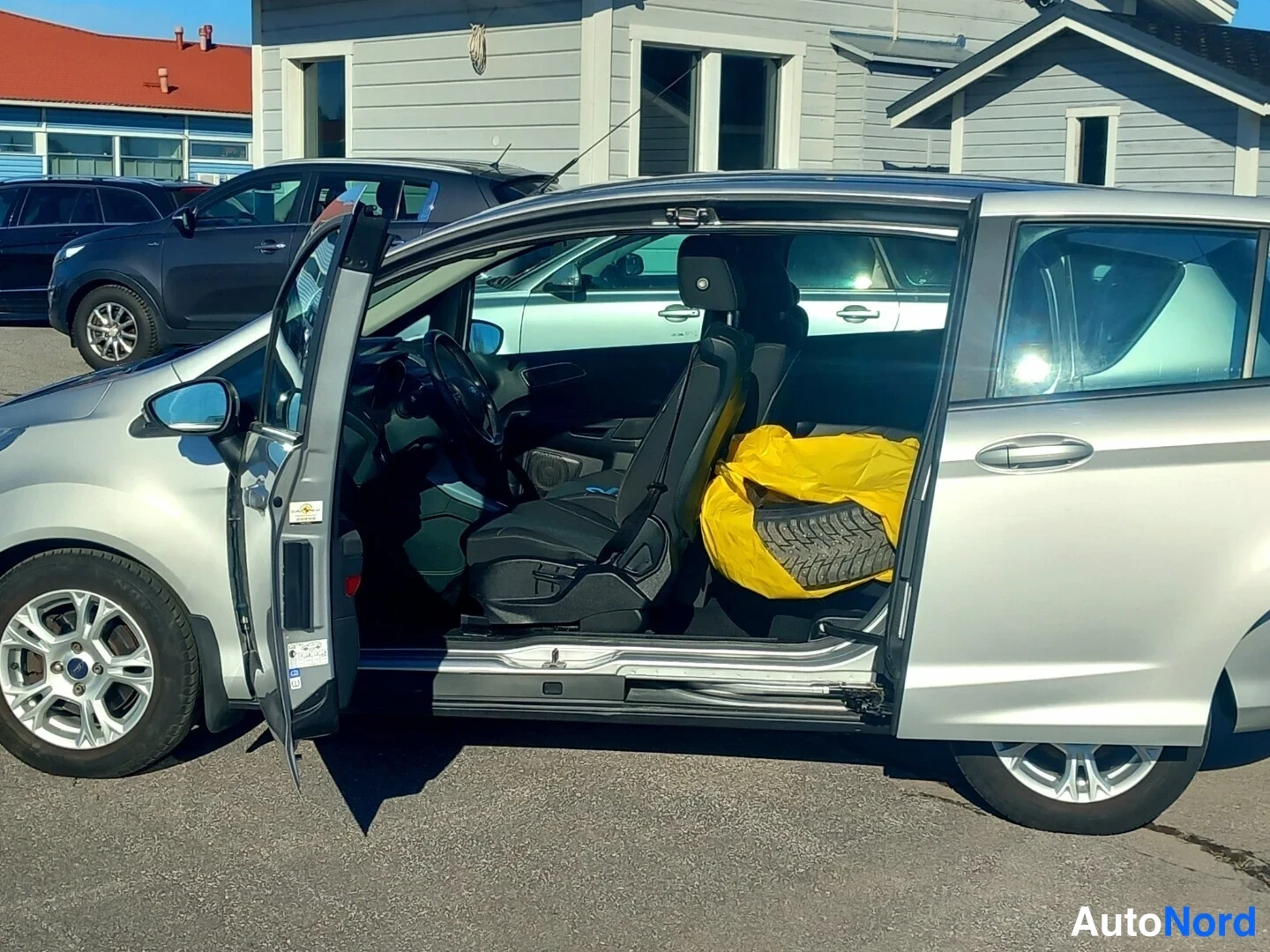 Sivulta: Hopeanvärinen auto sivusta, molemmat ovet auki; sisältä näkyvät etuistuin, ohjauspyörä ja keltaisen suojan alla oleva varaosa.