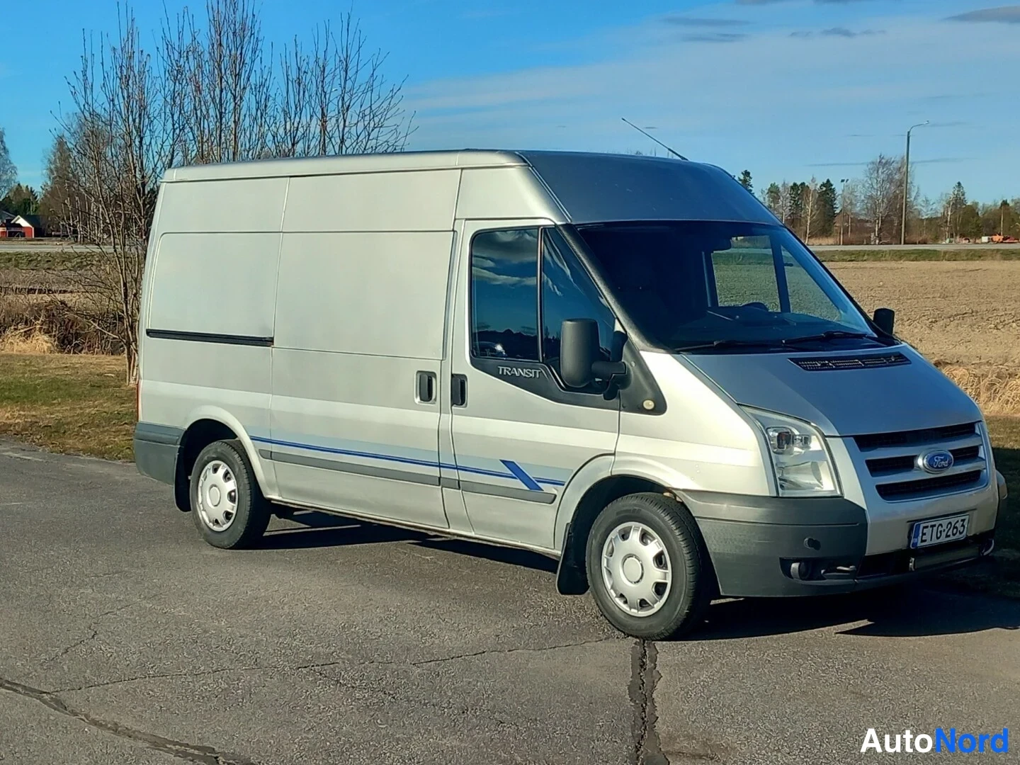 Etuviisto, nokka oikealle: Ford Transit -pakettiauto hopeanvärisenä kolmesta neljäsosasta etuosasta oikealta, etusäleikkö ja rekisterikilpi näkyvissä.