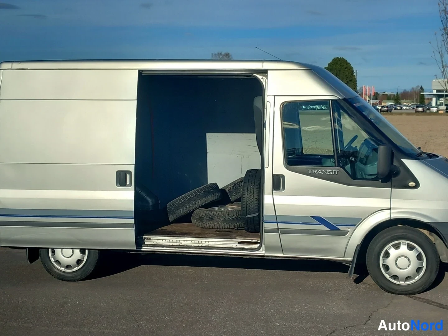 Sivulta: Hopeanvärinen Ford Transit -pakettiauto sivulta, liukuov auki ja renkaita lavalta näkyvissä.