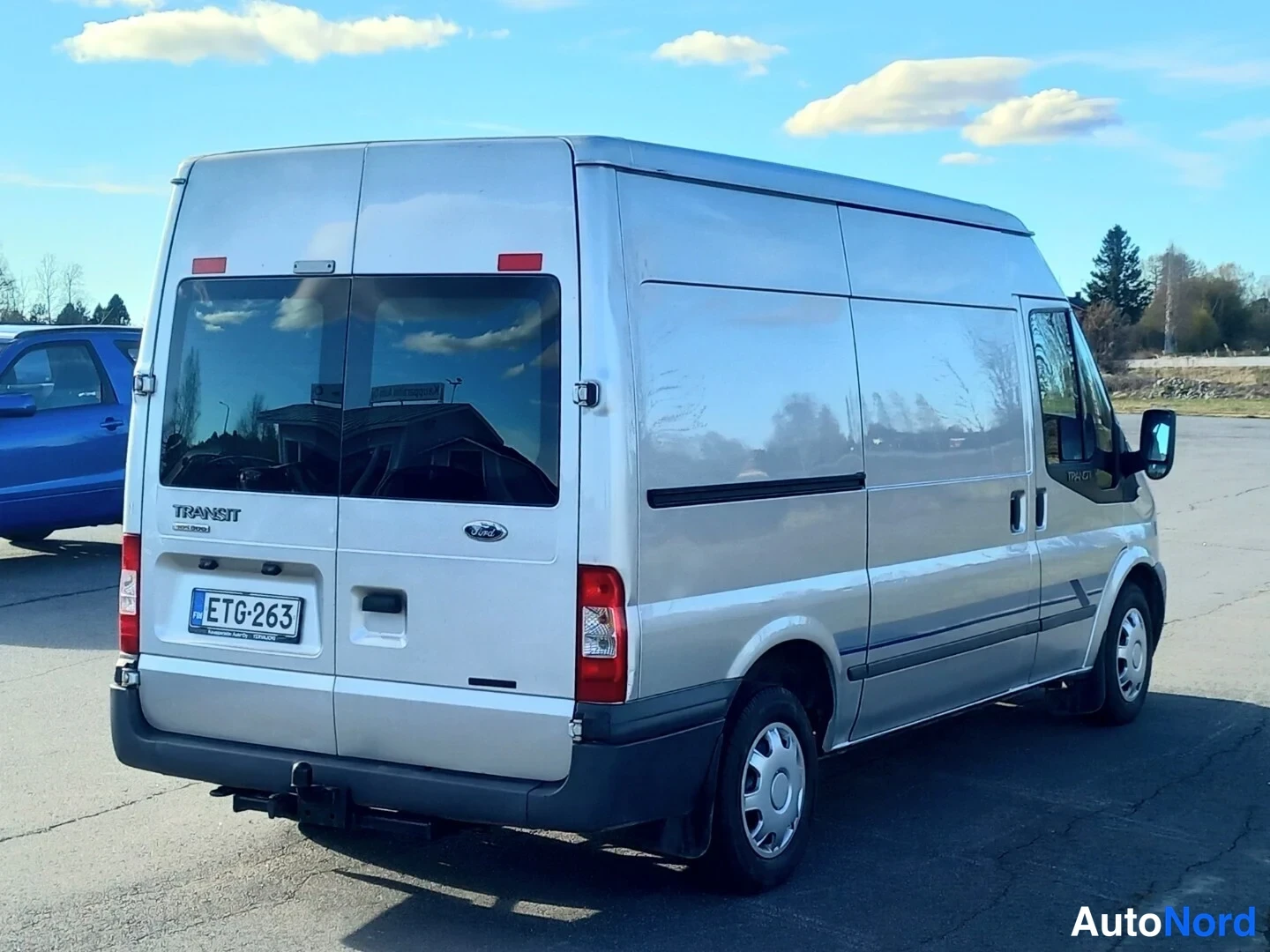 Takaviisto: Hopeanharmaa Ford Transit -pakettiauto takaa sivusuunnasta kuvattuna, rekisterikilpi, takana TOW BAR -kytkin ja Transit-merkintä.