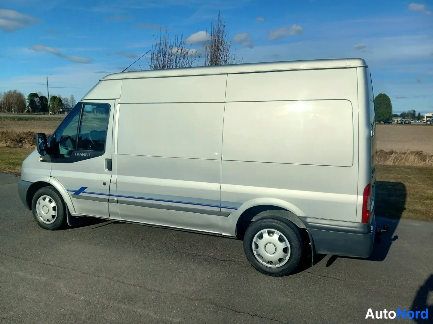 Takaviisto: Ford Transit -pakettiauto hopeanvärisenä sivulta takavasemmalta, koripaneelit ja sininen koristelinja näkyvissä.