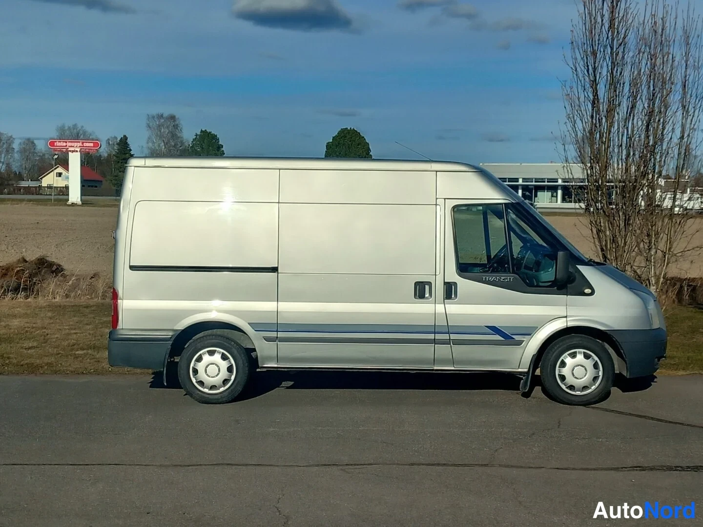 Sivulta: Hopeanvärinen pakettiauto sivulta oikealta, transit-teksti ikkunassa, sininen koristelista kyljessä.