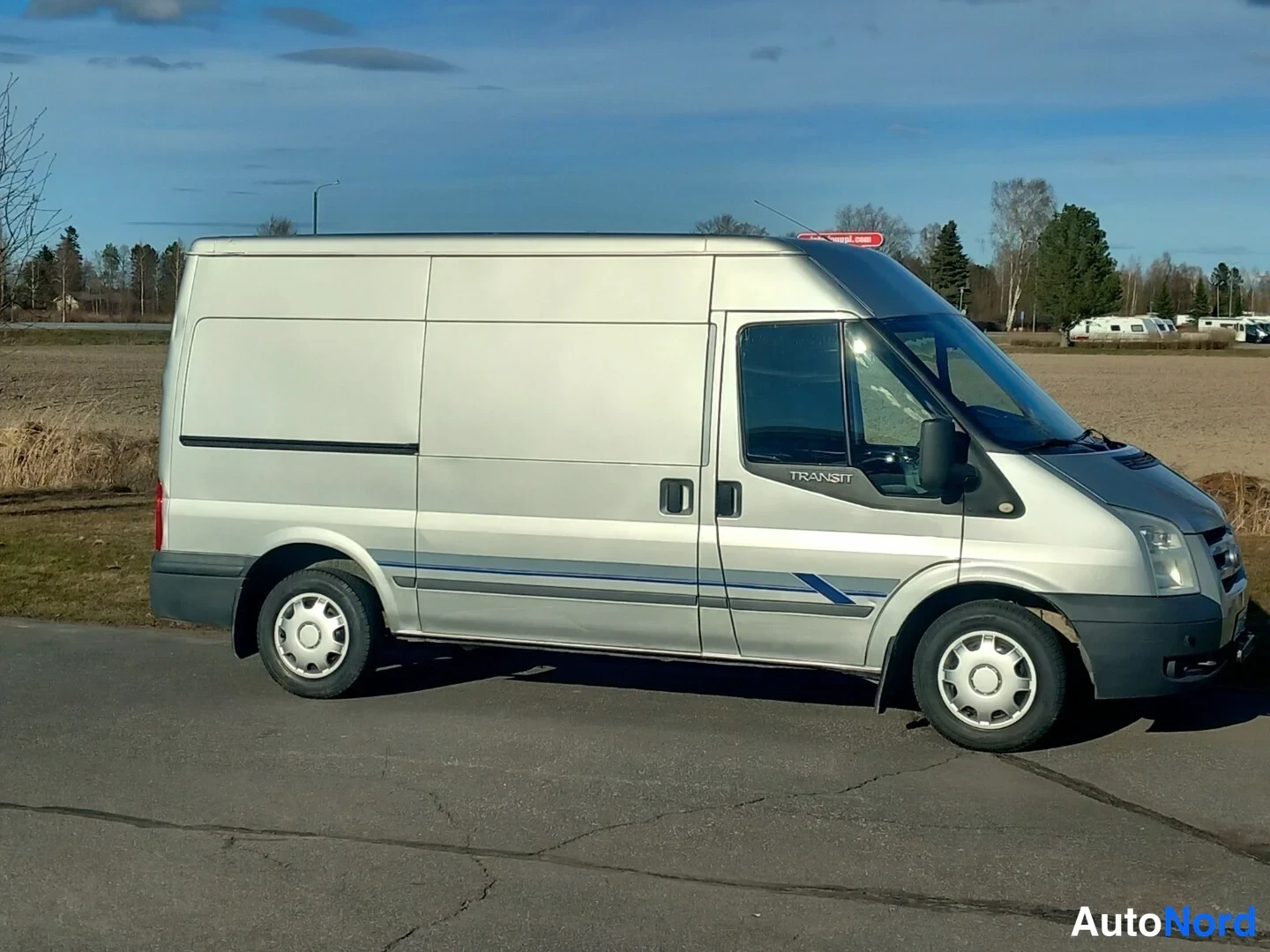 Etuviisto, nokka oikealle: Ford Transit -pakettiauto hopeanvärisenä sivulta etuosa näkyvissä asfaltilla.