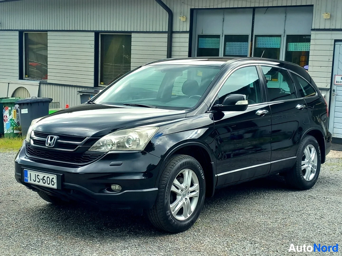 Etuviisto: Musta Honda CR-V -maastauto kolmesta neljänneskulmasta edestä vasemmalla, rekisterikilpi I JS-606, hopeavanteet, sora-asfaltti ja rakennus taustalla.