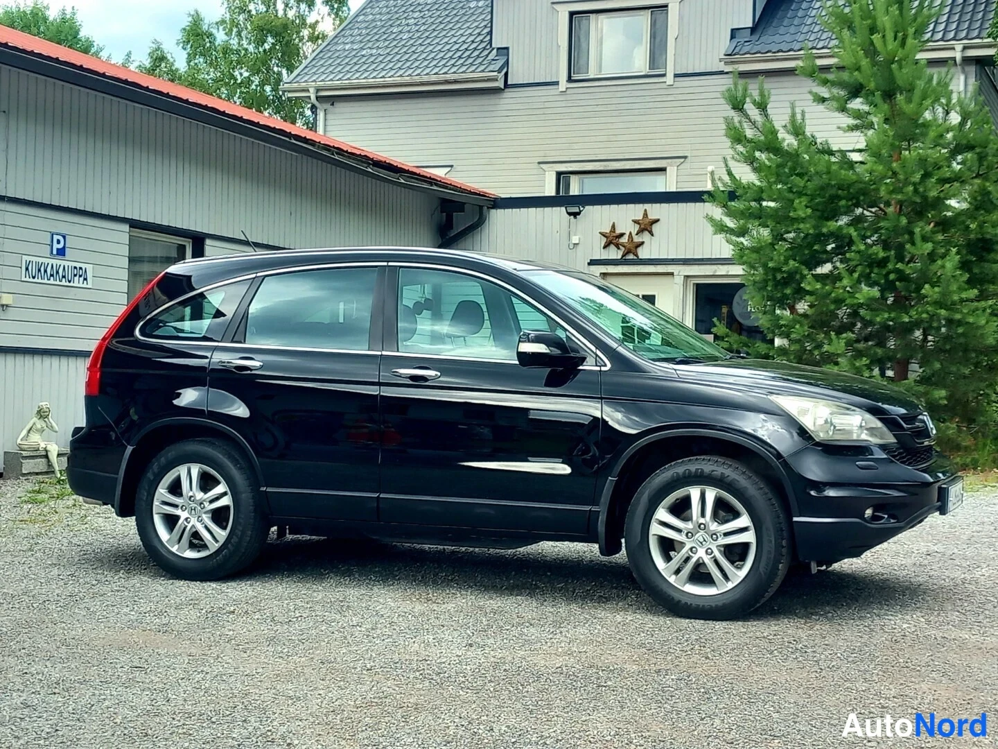 Etuviisto: Musta Honda CR-V etuviistosta oikealta, konepelti, ajovalot ja etupuskuri näkyvissä talon pihalla.