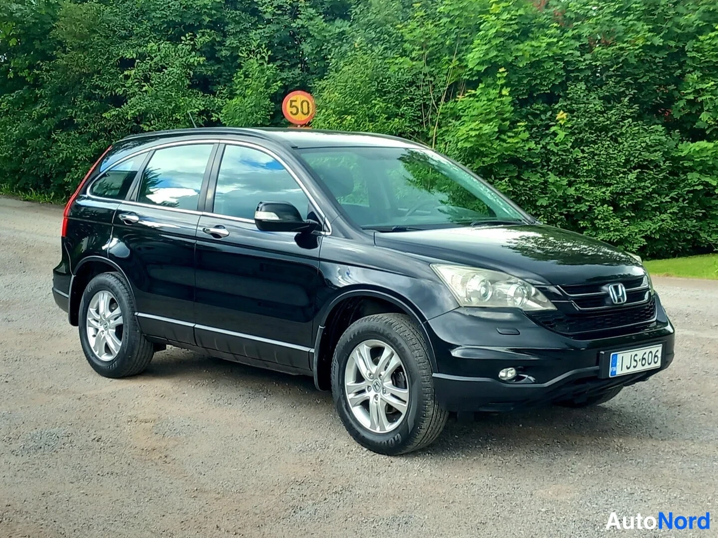 Etuviisto: Musta Honda CR-V kolmanneksesta oikealta edestä, rekisterikilpi I JS-606, takana avattu tavaratilan suu, 50-kilometrin nopeusrajoitustaulu taustalla, sora/asfal