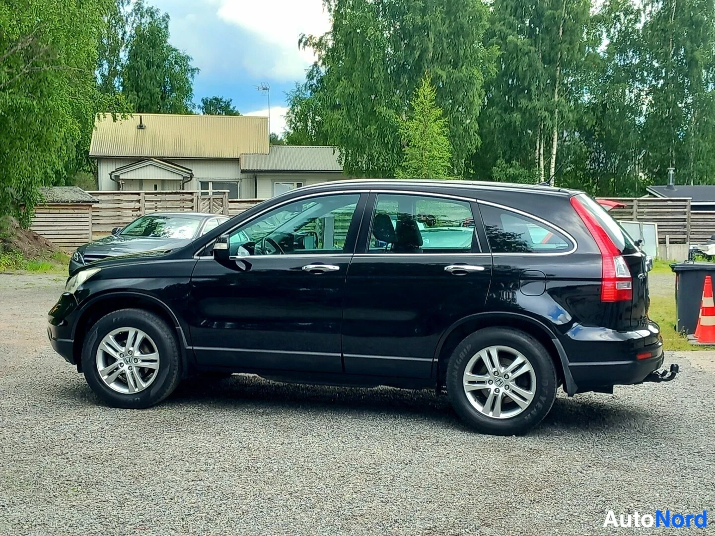 Sivulta: Musta Honda CR-V:n vasen sivuprofiili hiekkaisella pihalla, rakennukset ja puut taustalla.