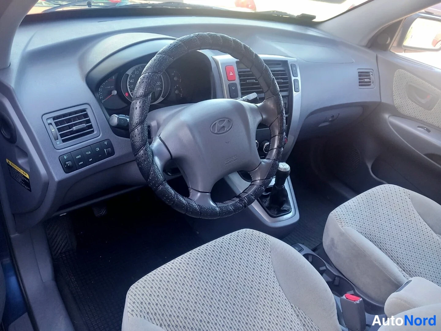 Muu: Hyundai-ohjauspyörä mustalla päällysteellä, mittaristo ja manuaalivaihtaja vaalean kankaan istuinten edessä.