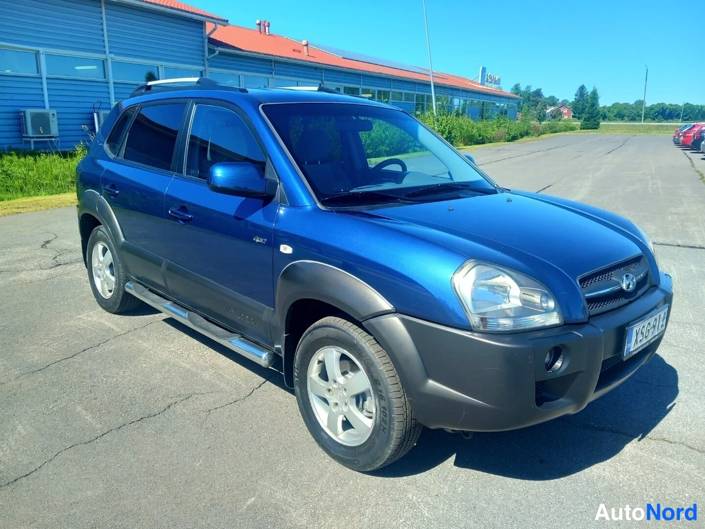 Etuviisto, nokka oikealle: Sininen Hyundai Tucson kulmassa kuvattuna etuosasta ja vasemmasta sivusta aurinkoisella parkkipaikalla.