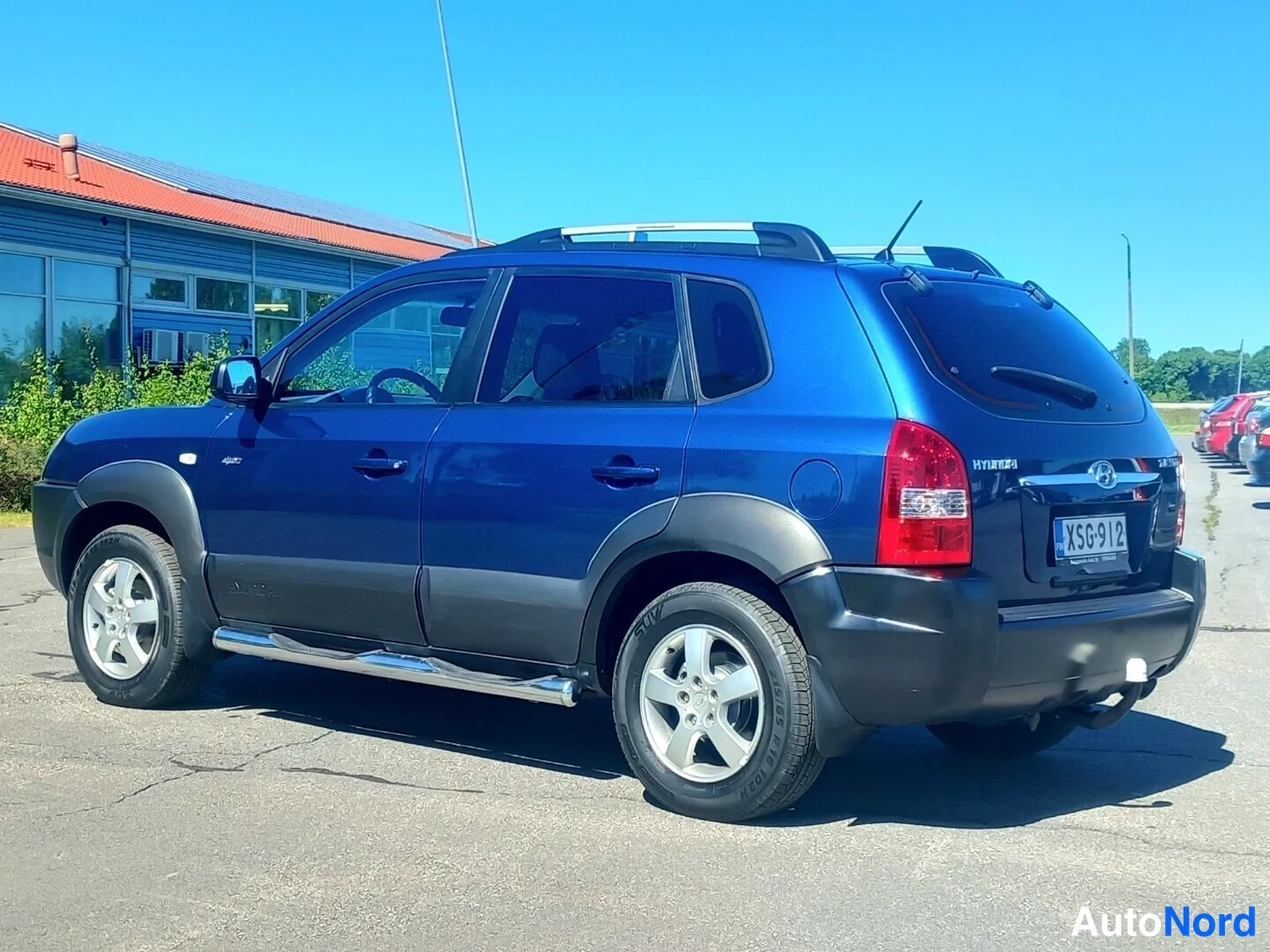 Takaviisto: Sininen Hyundai Tucson takaa ja vasemmalta sivulta kuvattuna; näkyvissä tumma takapuskuri, kattokaiteet, sivulaudat ja takanäyttö