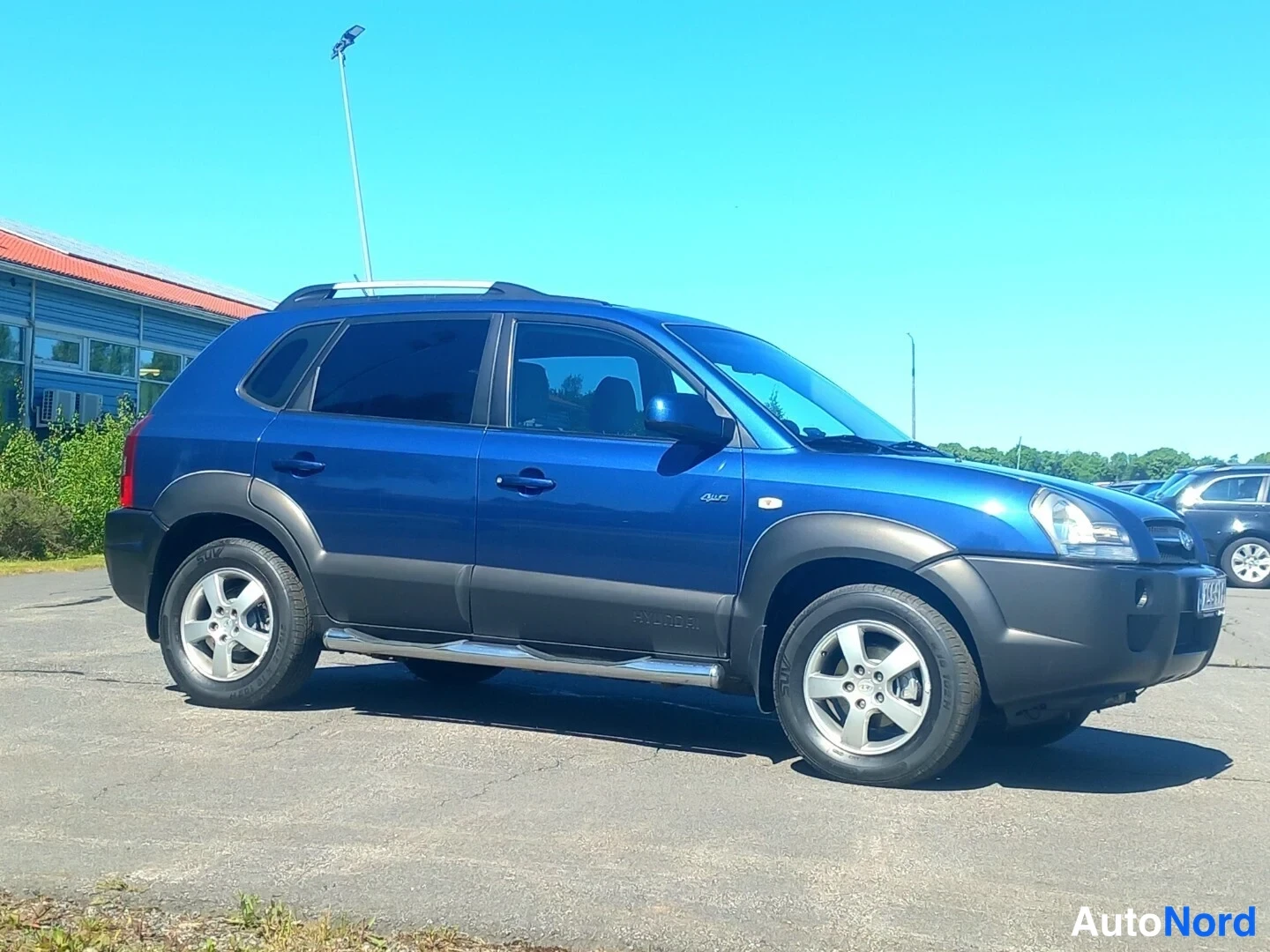 Etuviisto, nokka oikealle: Sininen Hyundai Tucson -urheilumaasturista etukulmakuva, jossa näkyvät kattokaiteet, kylkiskaiteet ja viisipuolaiset alumiinivanteet auringonpaisteessa p