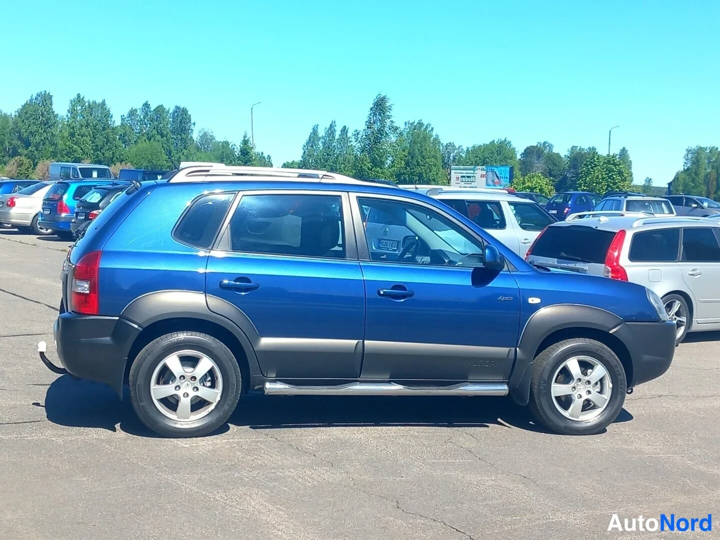 Takaviisto: Sininen Hyundai Tucson takaa vasemmalta sivulta kuvattuna; perävaunukenkä, kattokaiteet ja sivukaiteet näkyvät.