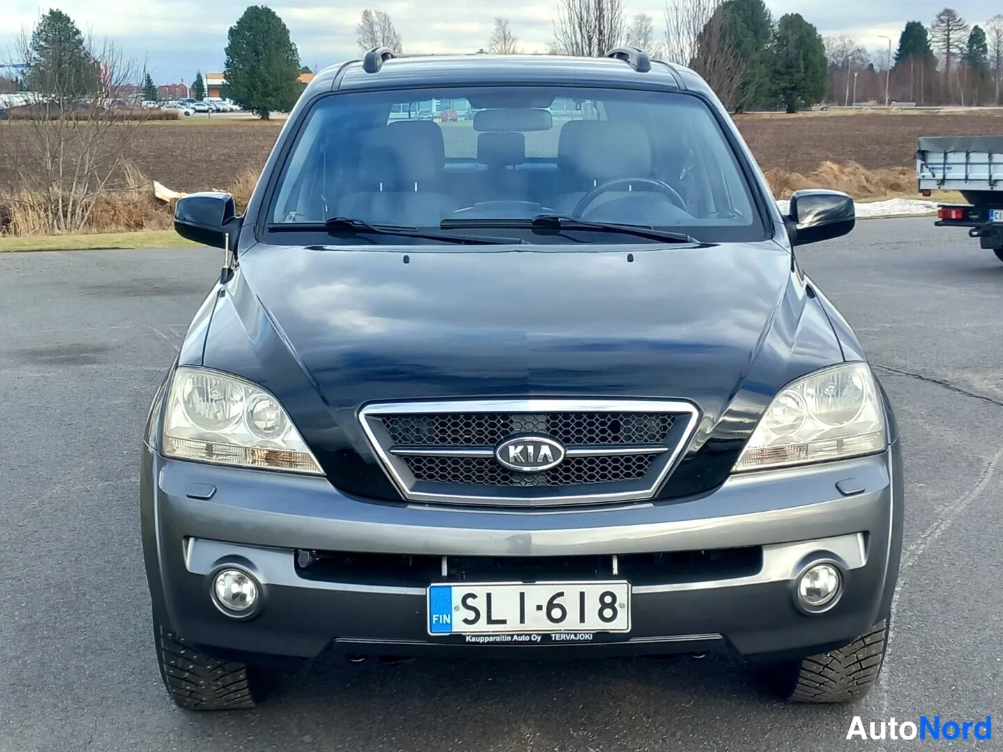 Edestä: Kia Sorenton etunäkymä suoraan edestä; musta konepelti, harmaa puskuri, KIA-merkki sälepäteissä, sumuvalot ja rekisterikilpi.