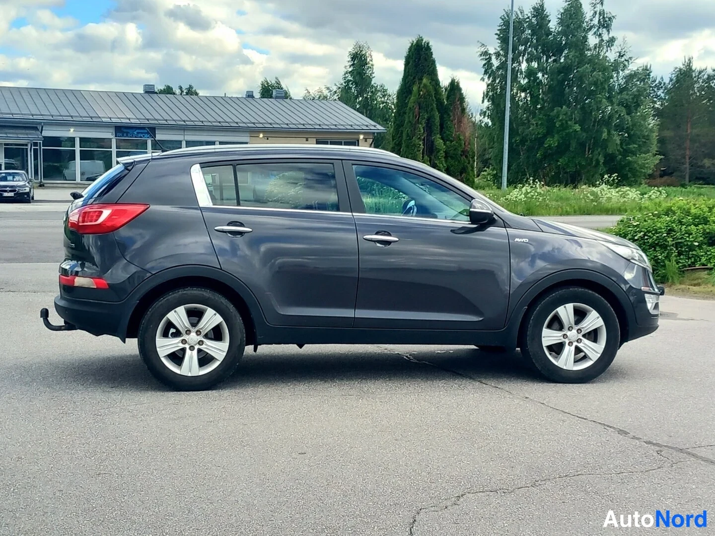 Takaviisto: Kia Sportage takaa oikealta kuvattuna; takavalot ja tavaratilan luukku sekä oikea kylki näkyvissä, autotalli ja muut ajoneuvot taustalla.