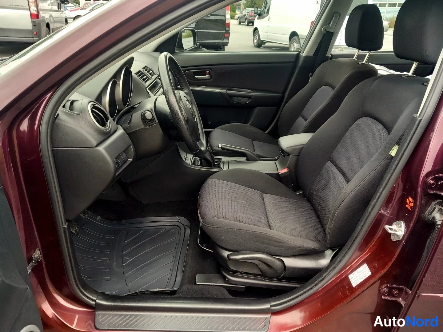 Muu: Auton etumatkustustila ovella katsottuna: kaksi tummaa kangasistuinta, Mazda-ohjauspyörä ja kumimatot.