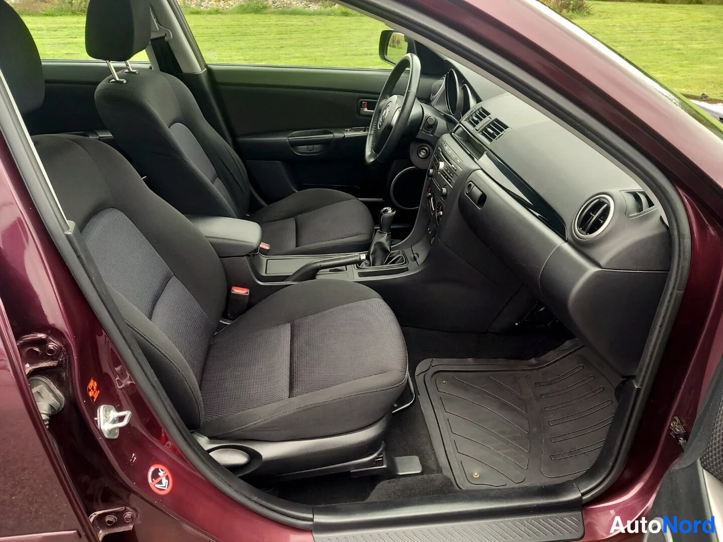 Muu: Auton sisätilat takaa eteenpäin: tummat kangasistuimet, Mazda-ohjauspyörä, manuaalivaihteisto ja kumimatto.