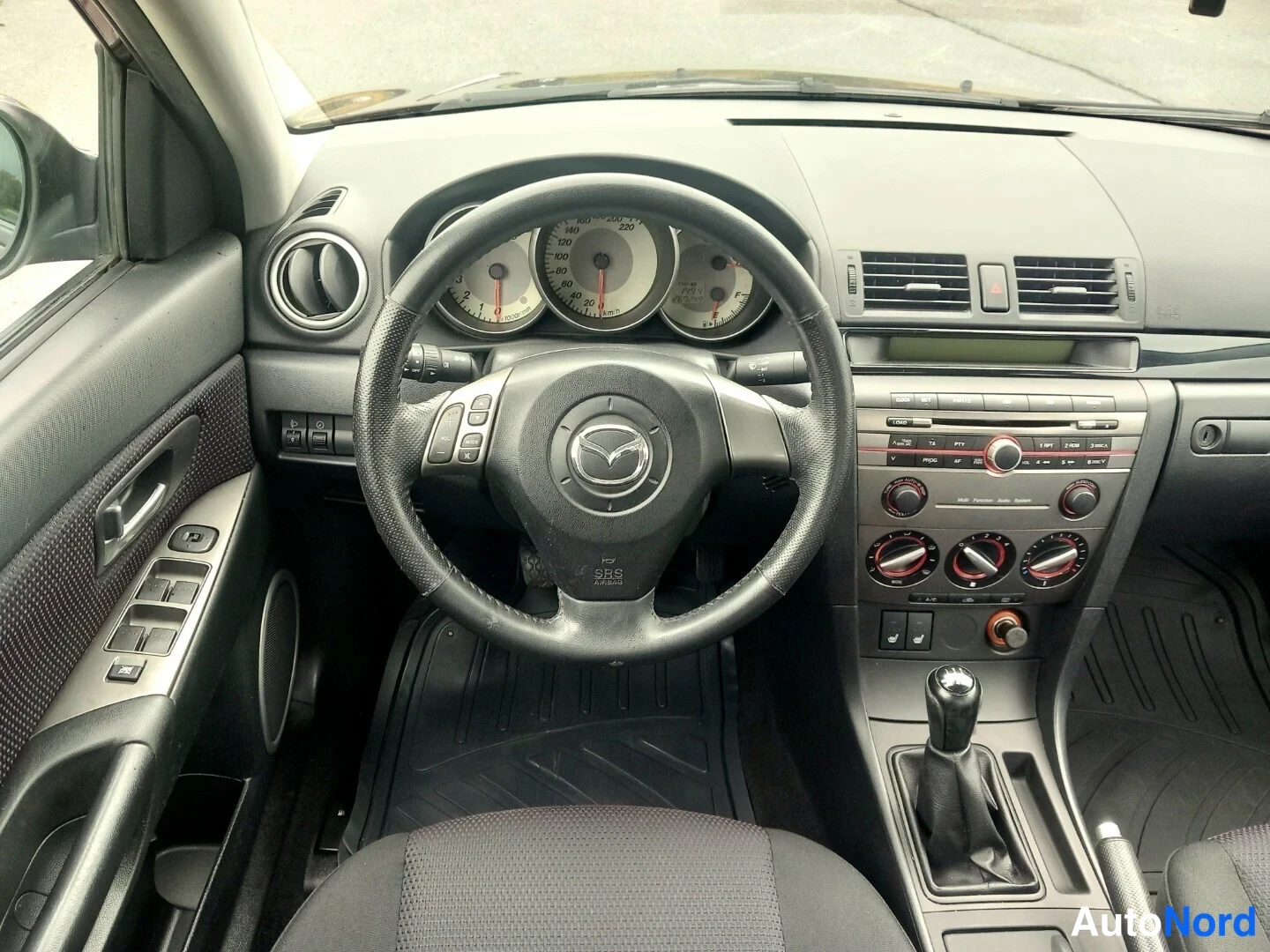 Muu: Ohjaamo Mazda-merkillä: rattivakokoneisto, manuaali, musta kojelauta ja vaihdin.