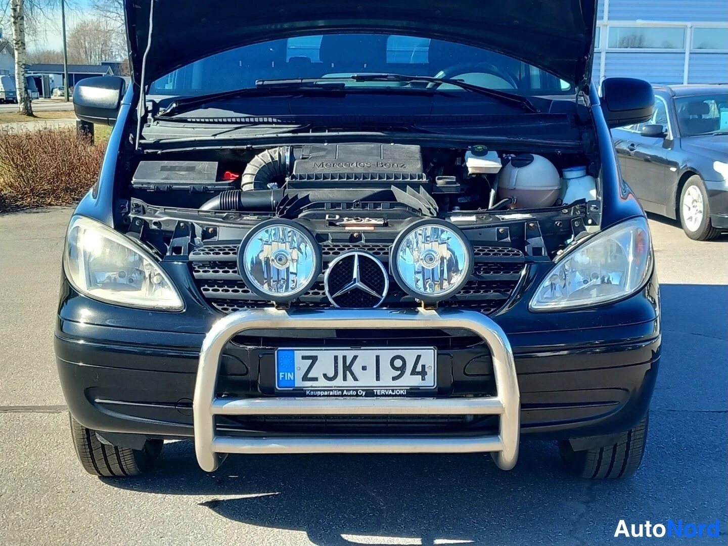 Edestä: Auki nostetun konepellin alla mustan Mercedes-Benz Vito -auton moottoritila, Mercedes Benz -teksti moottorin kannessa, etupuskurin suojakaari ja rekisterikilpi.