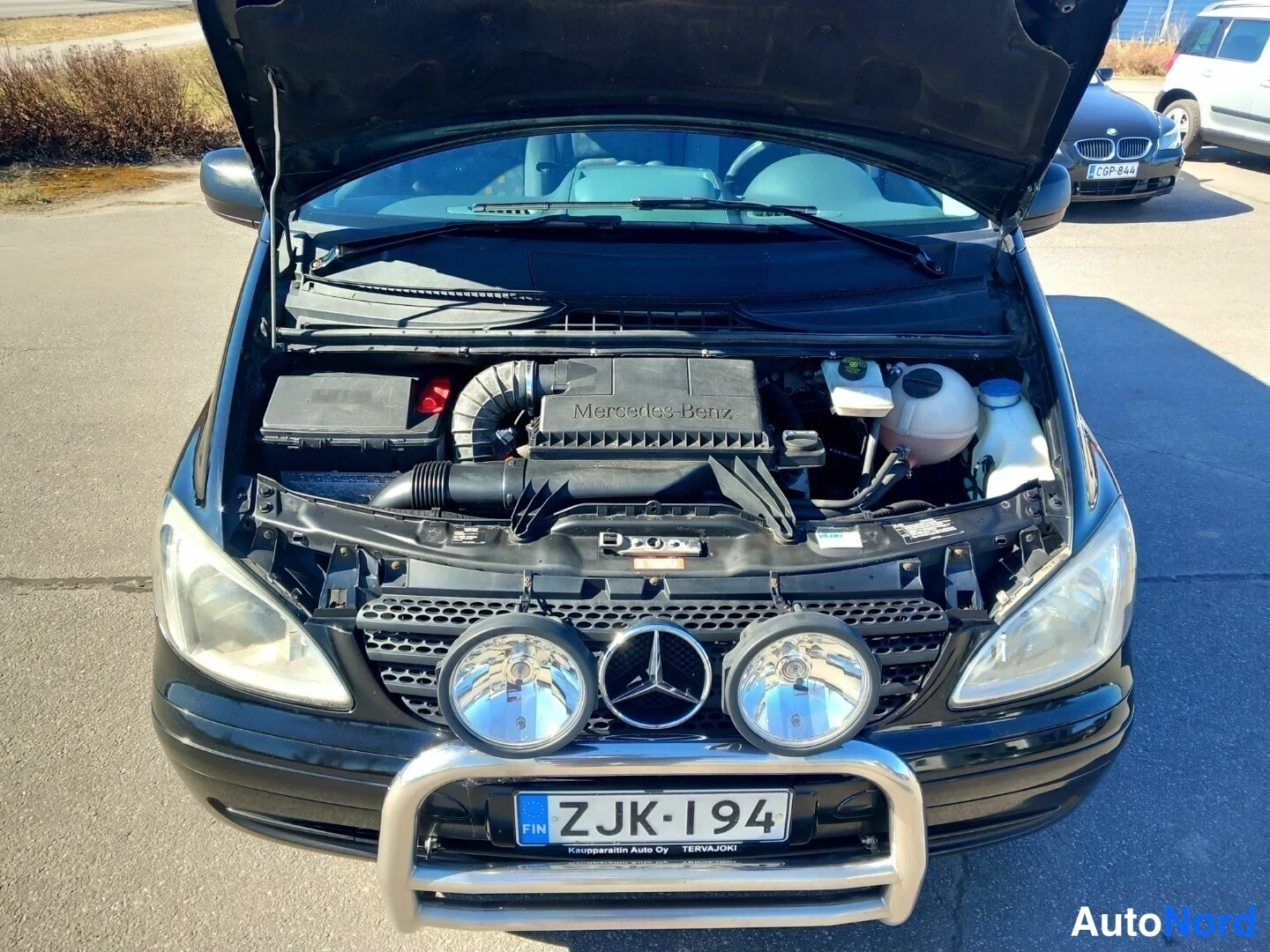 Edestä: Moottoritila avattuna, Mercedes-Benz-merkintä moottorin kannessa, etupuskurissa kaksi lisävaloa ja suojakaari, rekisterikilpi.