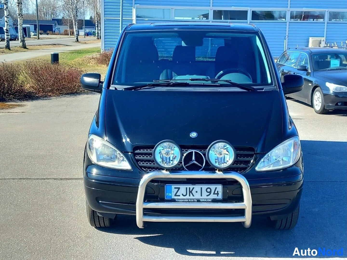 Edestä: Mustan pakettiauton keula edestä: kaksi lisävaloa ja metallinen keulakaari rekisterinumeron yllä.