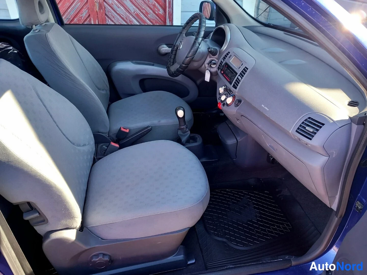 Muu: Auton sisätilat matkustajan puolelta katsottuna: kaksi harmaata kangastuolia, Nissan-ohjauspyörä mustalla päällillä, keskikonsolin säätimet, radio ja mustat mat