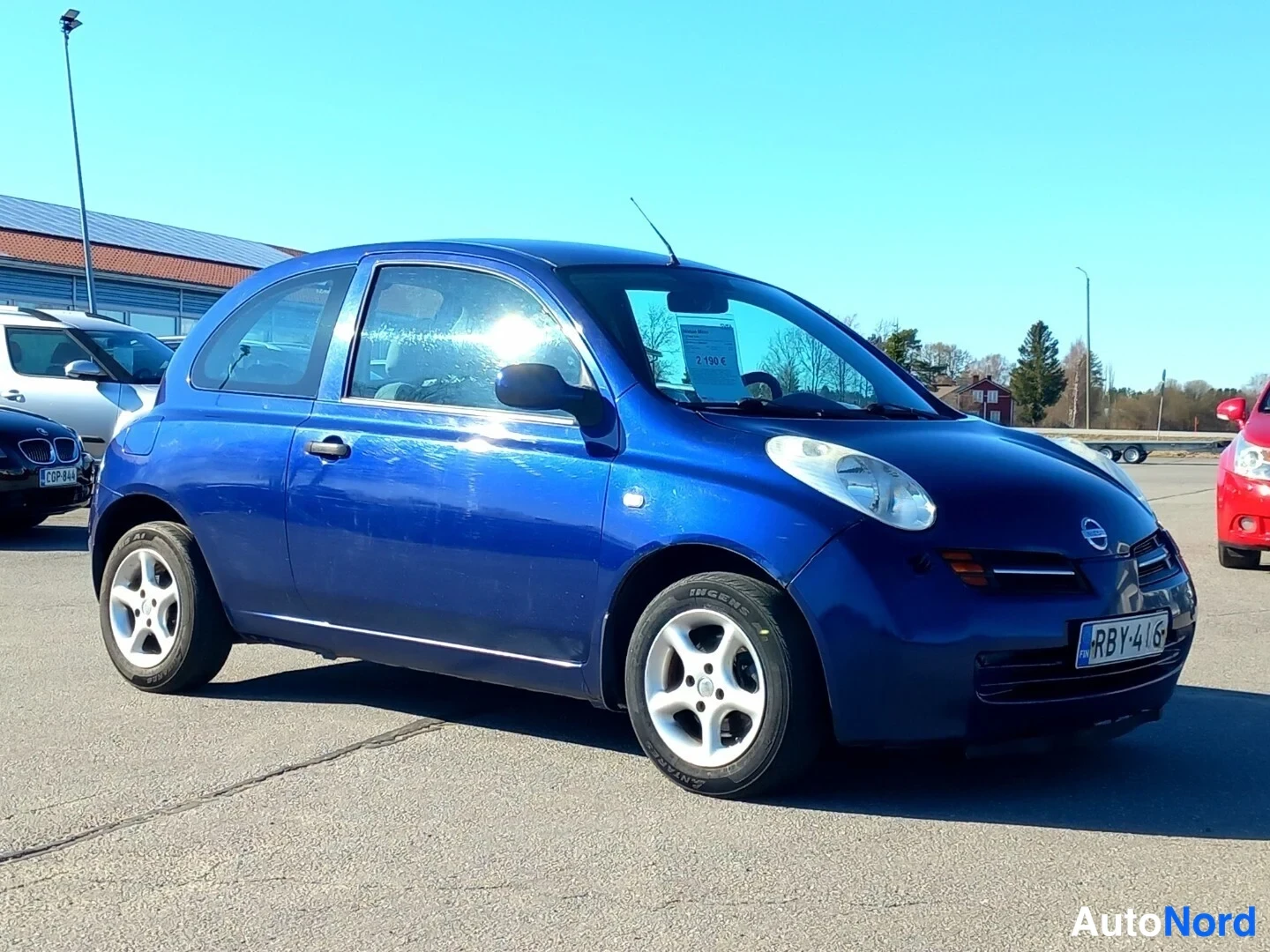 Etuviisto, nokka vasemmalle: Sininen Nissan Micra edestä vasemmalta kuvattuna auringonpaisteessa, tuulilasissa keltainen hinnoittelulappu.