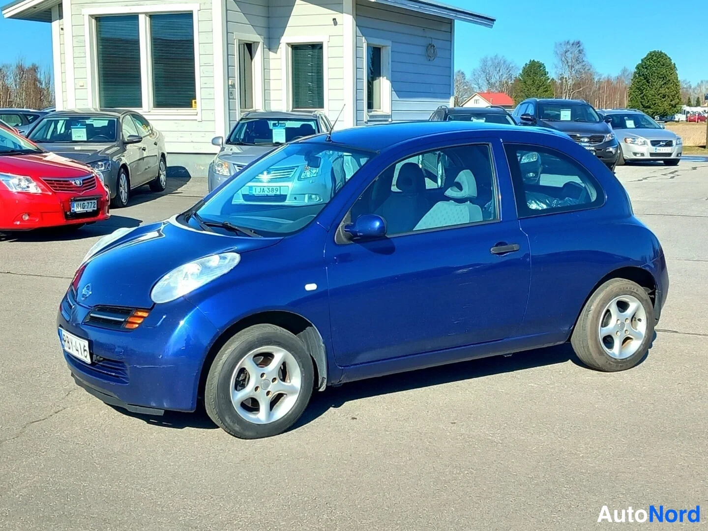 Etuviisto, nokka vasemmalle: Sininen Nissan Micra edestä vasemmalta kuvattuna autopaikalla.