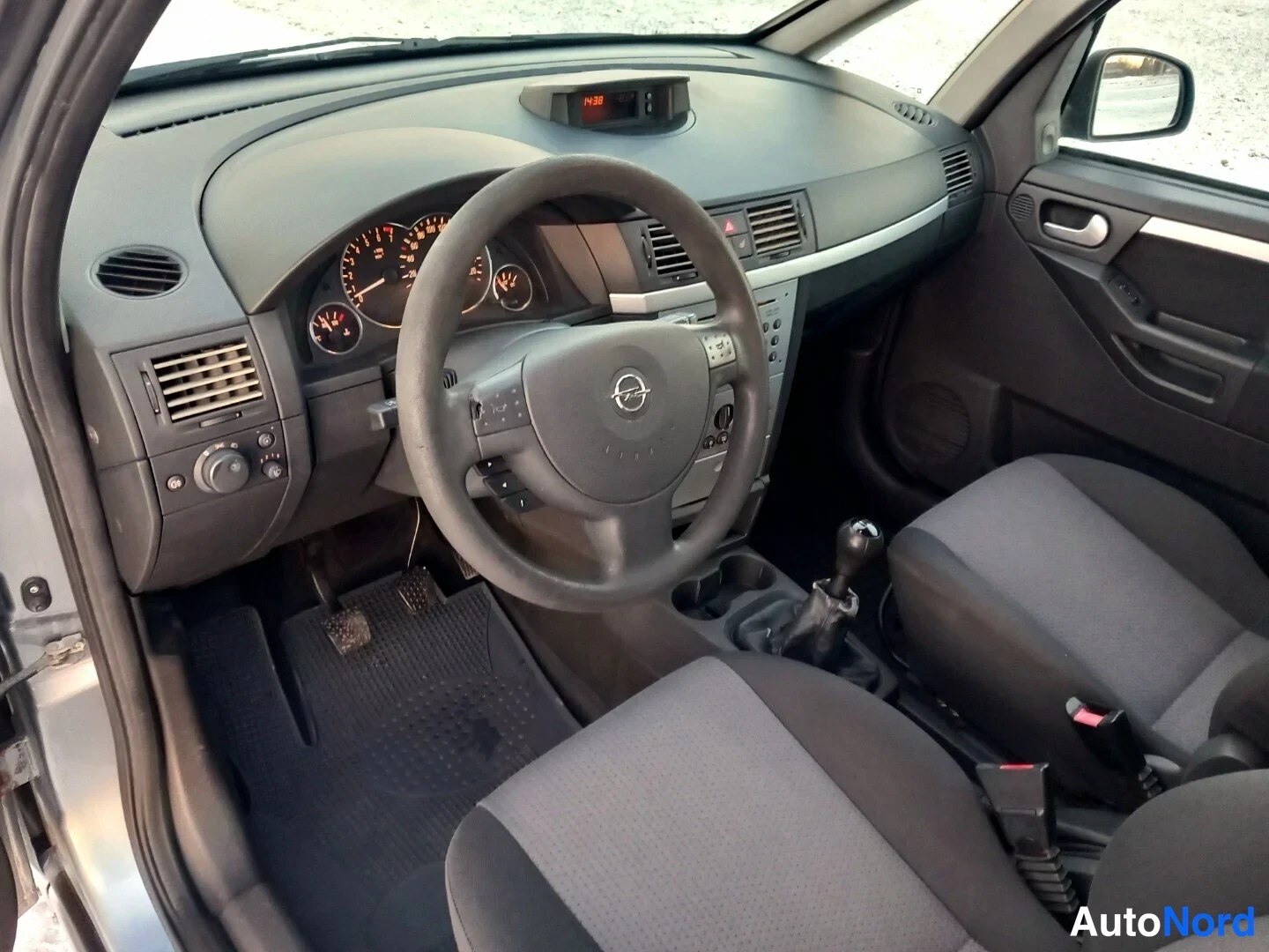 Muu: Sama ohjaamon näkymä toista kulmaa: Opel-ohjauspyörä, mittaristo, manuaalivaihde ja tummat istuimet.