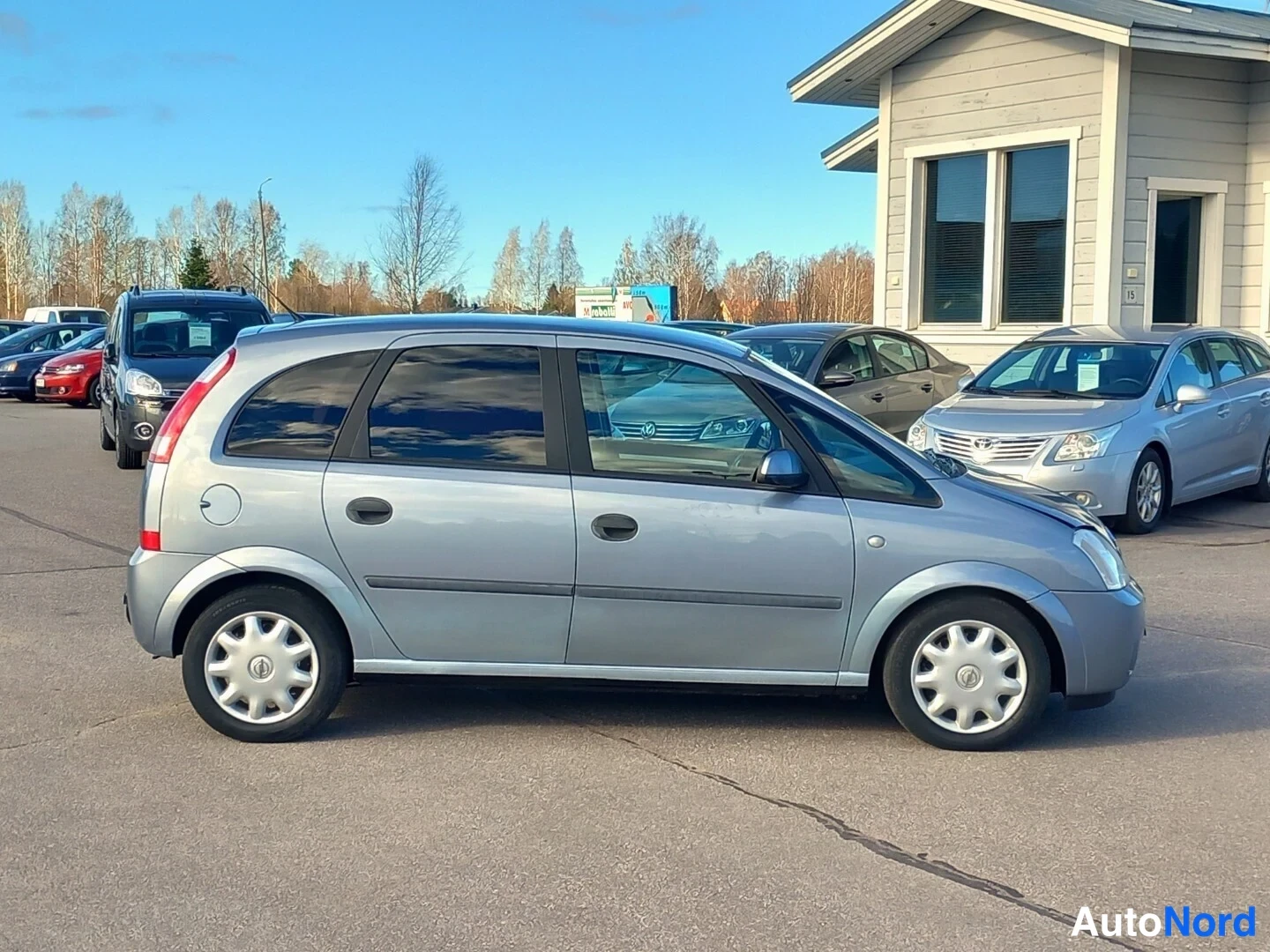 Etuviisto, nokka oikealle: Opel Meriva -auton sivukuva oikealta puolelta, etupaneelin kulma ja oikea kylki näkyvissä, hopeinen koriväri.