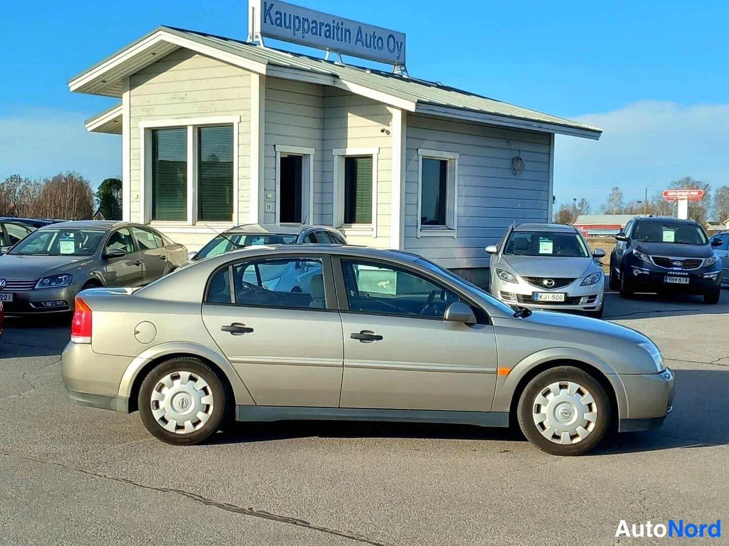 Takaviisto: Beige Opel Vectra -sedan takakulmasta vasemmalle päin autoliikkeen pihalla.