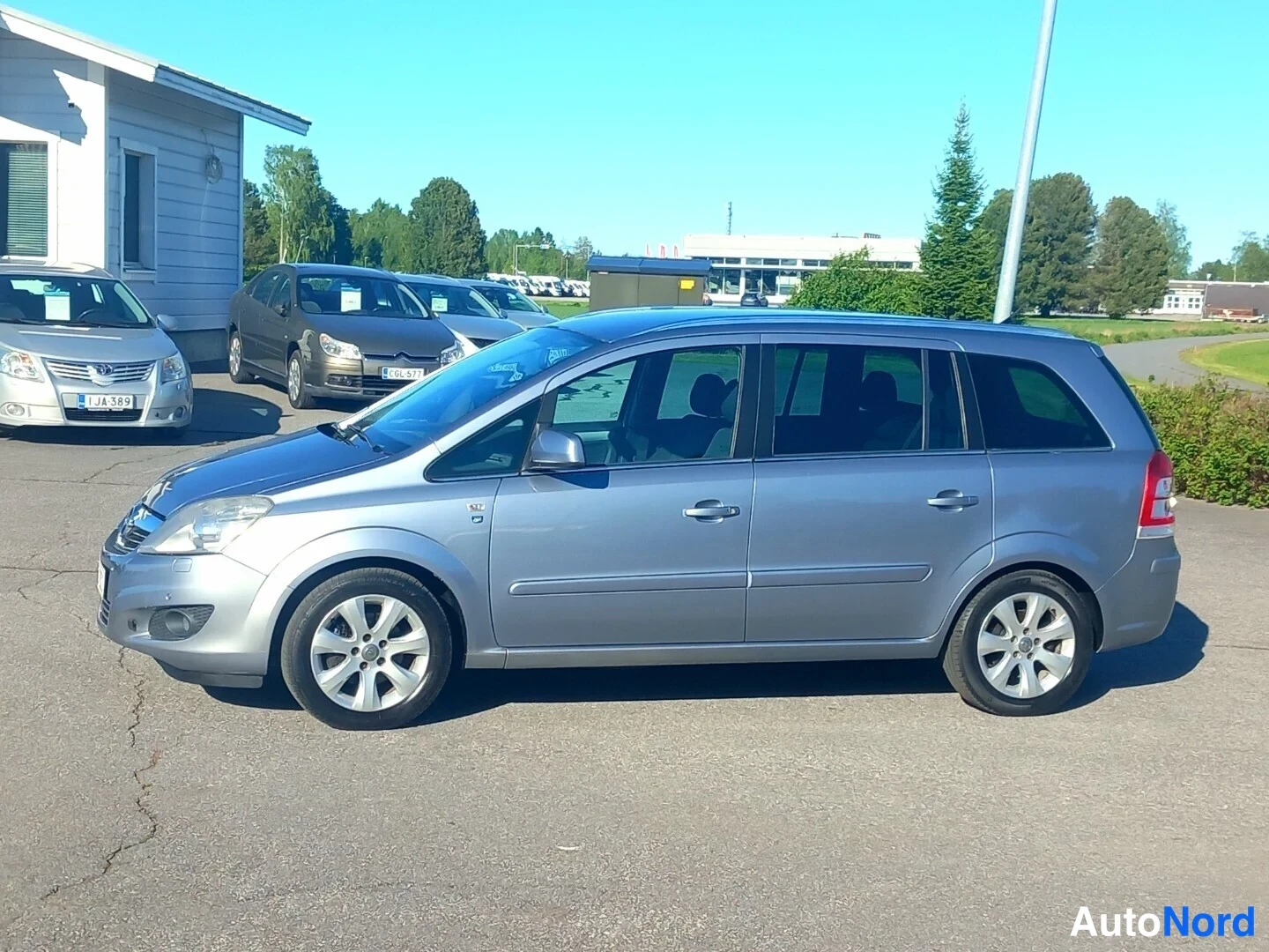 Etuviisto, nokka vasemmalle: Hopeanharmaa tila-auto sivulta etukulmasta, nokka vasemmalla, pihamaalla.