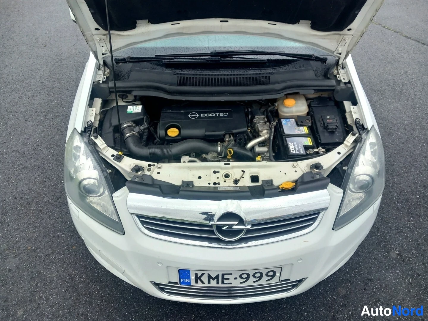 Edestä: Valkoinen Opel Zafira, konepelti auki, moottoritila ja etusäleikkö näkyviltä ylhäältäpäin.