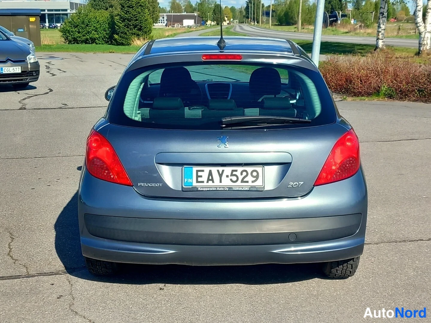Takaa: Suora takakuva hopeanharmaasta Peugeot 207 -hatchbackista asfaltilla päivänvalossa. Takavalot ovat kirkkaanpunaiset ja rekisterikilpi lukee. Tavaratilan katossa