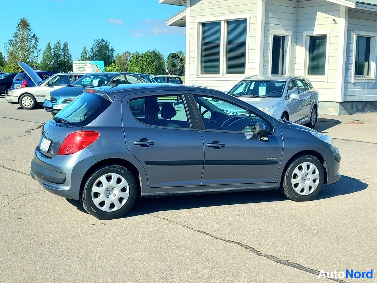 Takaviisto: Harmaa viistoperä Peugeot, takakolmannesvasen kuvakulma, auto parkkipaikalla muiden autojen edessä.