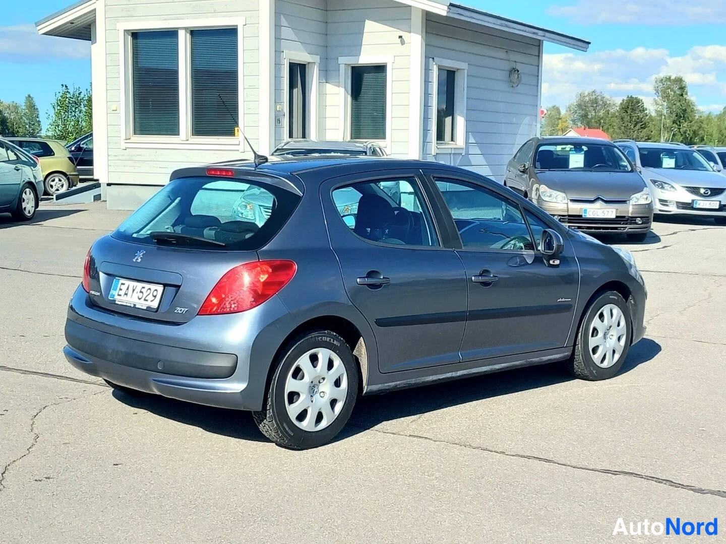 Takaviisto: Harmaa viisiovinen Peugeot 207 -porrashatchback takaa ja sivulta päin parkkipaikalla.