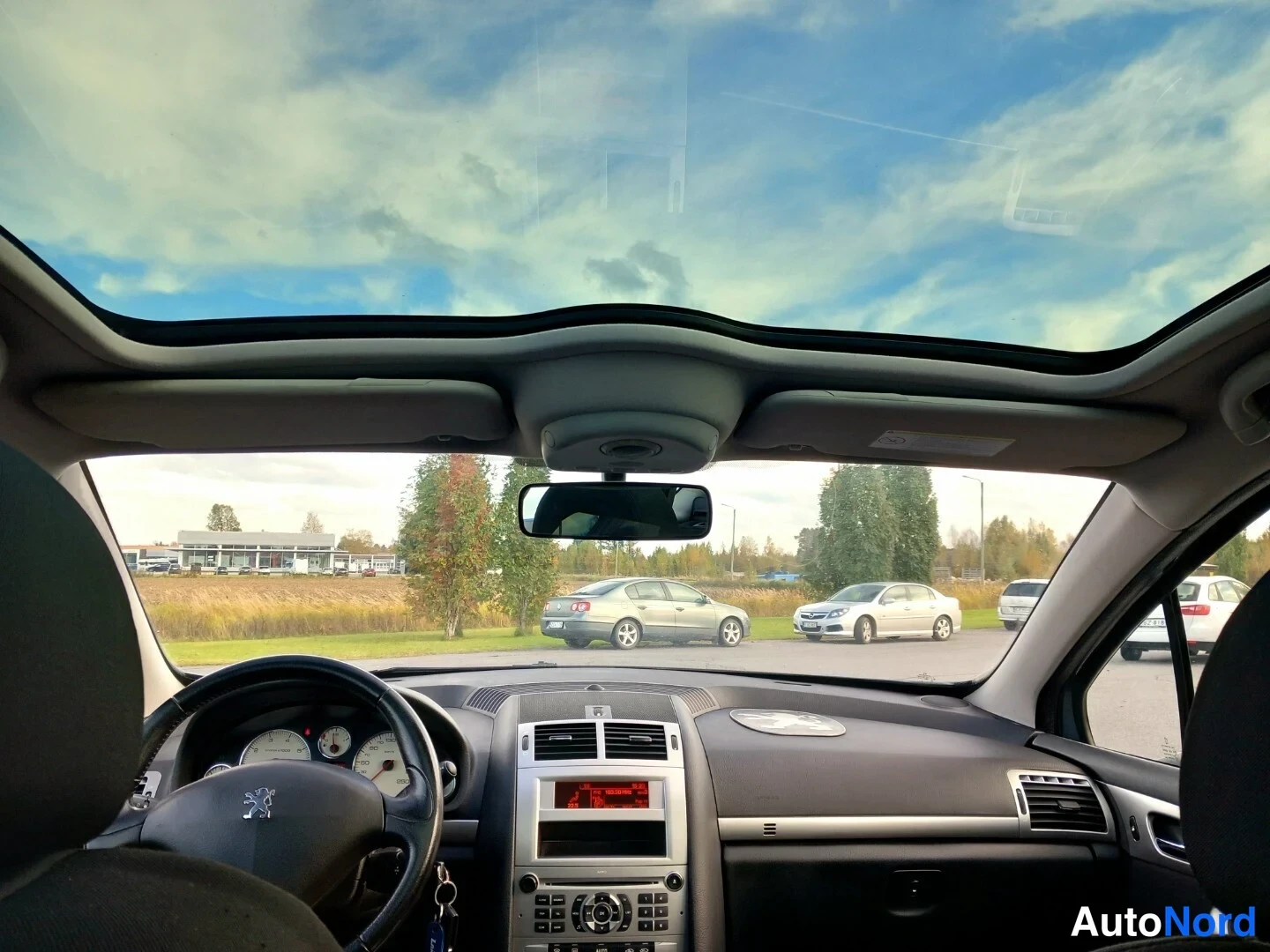 Muu: Näkymä auton takaosasta eteenpäin: panoraamalasikatto, Peugeot-merkkinen ohjauspyörä, mittaristo ja kojetaulu; taustalla parkkipaikalla muita autoja.