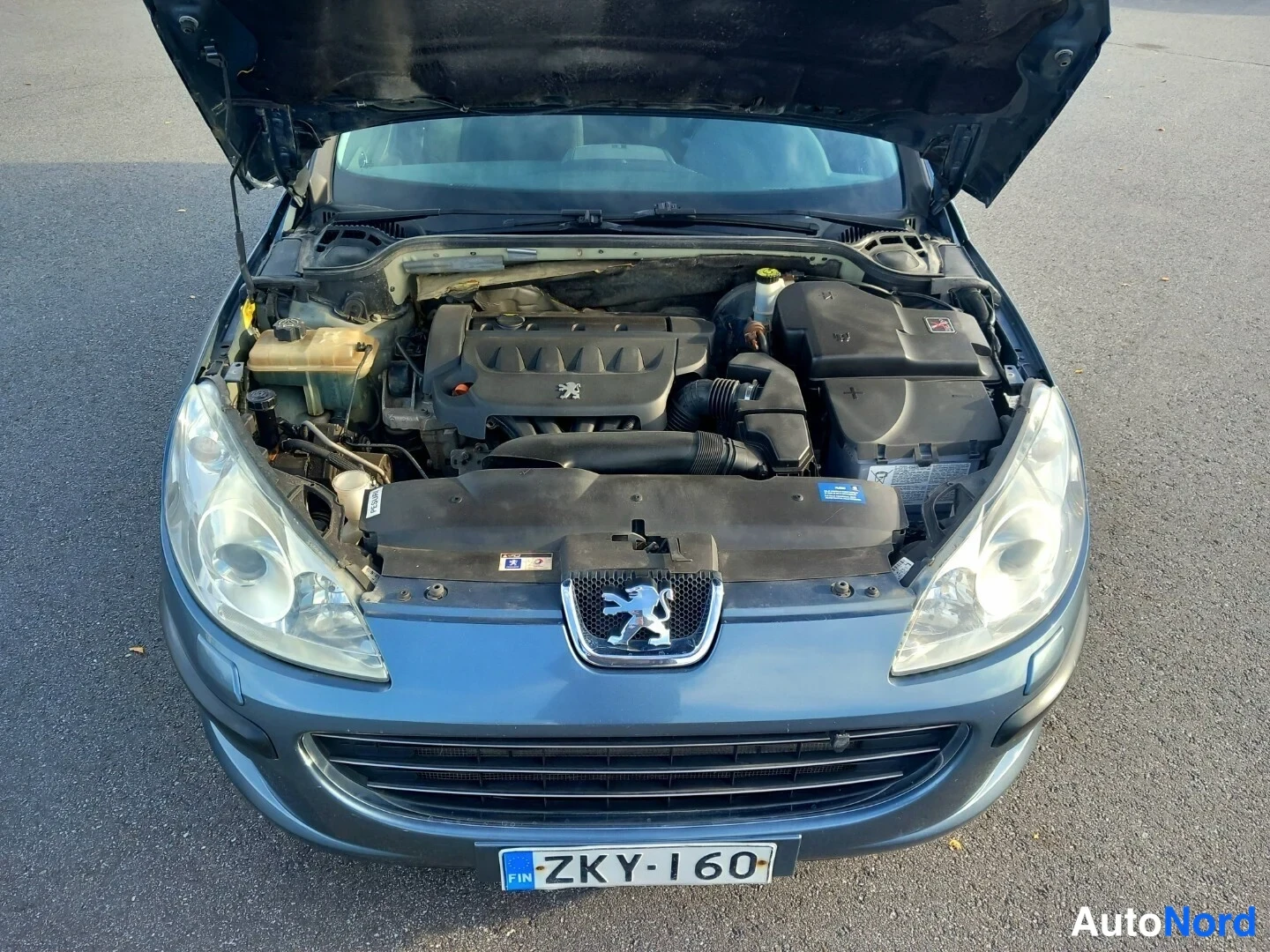 Edestä: Sinertävän harmaa Peugeot 407 edestä, konepelti auki ja moottoritila näkyvissä; rekisterikilpi.