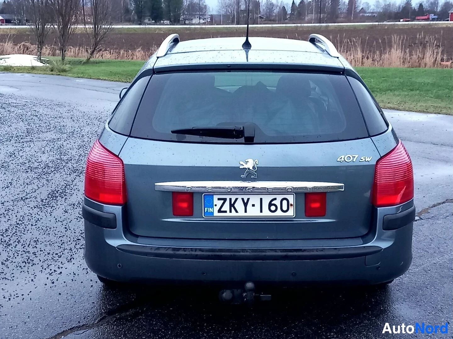 Takaa: Peugeot 407 SW:n takanäky; rekisterikilpi ja vetokoukun pallo, harmaa koriväri sateella märällä tiellä.