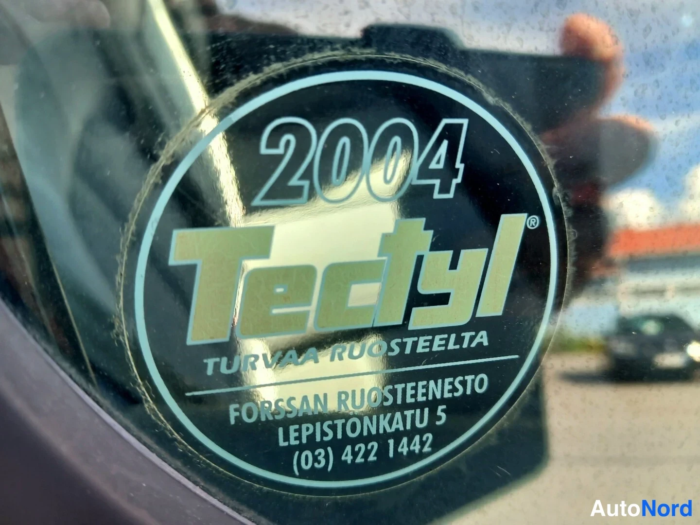 Muu: Tarkennettu kuva tuulilasin tarra: pyöreä Tectyl-tarra vuodella 2004 ja teksti 'TURVAA RUOSTEELTA' sekä Forssan Ruosteenson tiedot.