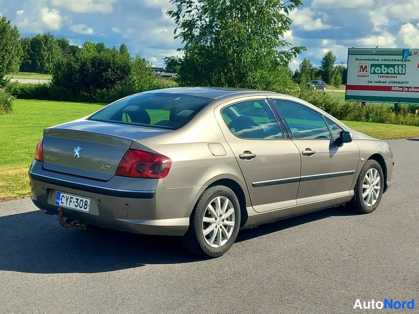 Takaviisto: Kultaisen Peugeot 407 -sedanin takaoikea kolmasosakuva. Auto seisoo asfalilla vihreän nurmikon edessä, ja taustalla näkyy mainostaulu.