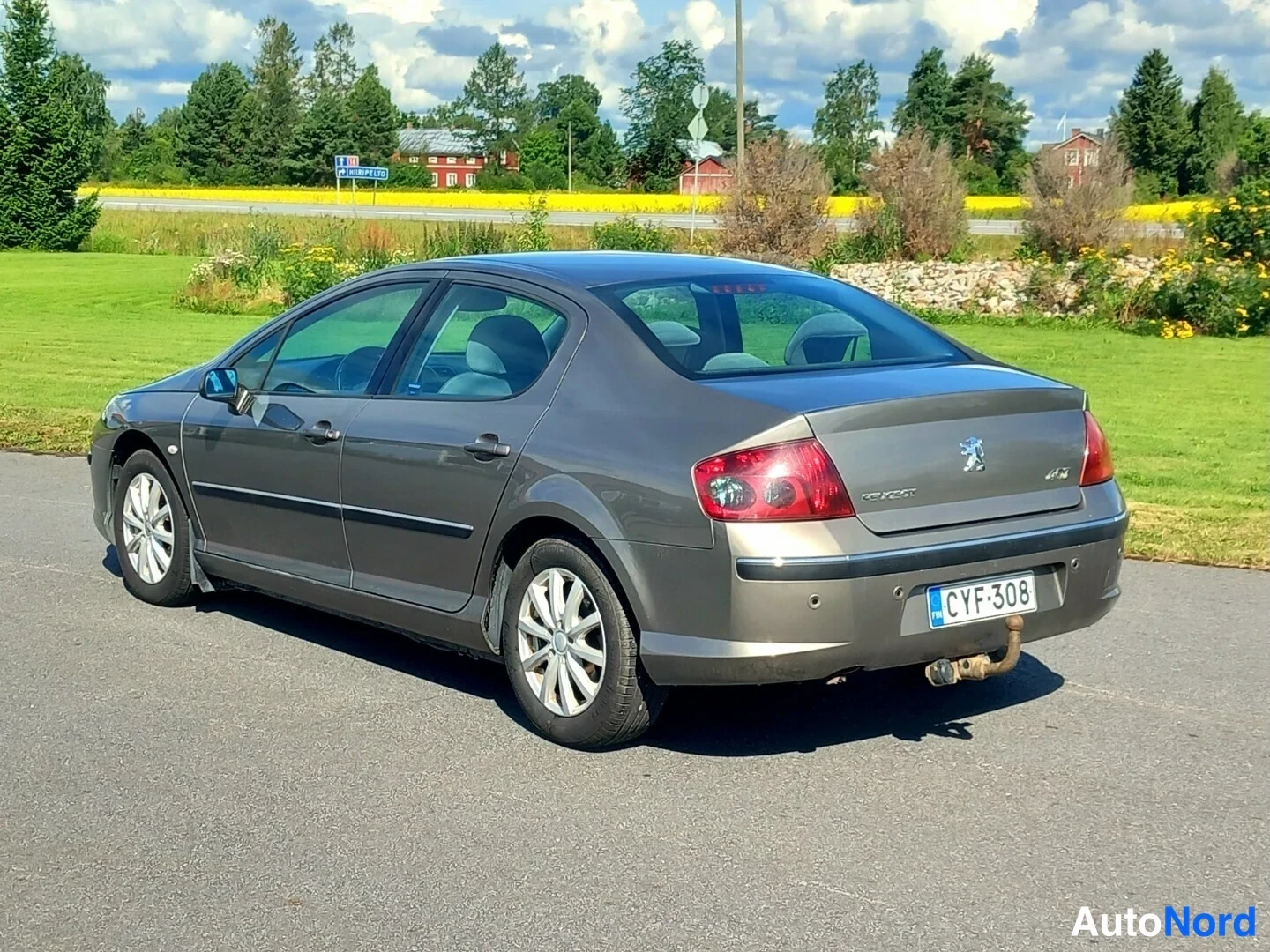 Takaviisto: Harmaan Peugeot 407 -sedanin vasen takaoikea kolmasosakuva. Auto seisoo asfaltille nurmikon reunalla, ja taustalla kukkivat keltaiset kasvit sekä punaisia taloj