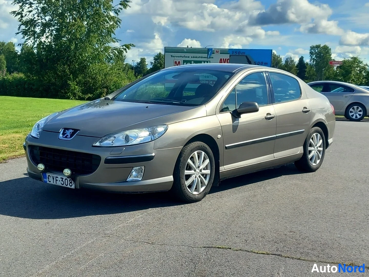 Etuviisto, nokka vasemmalle: Ruskeaharmaa sedan-tyyppinen henkilöauto kolmesivukulmassa, keulakko vasemmalla, rekisterikilpi.