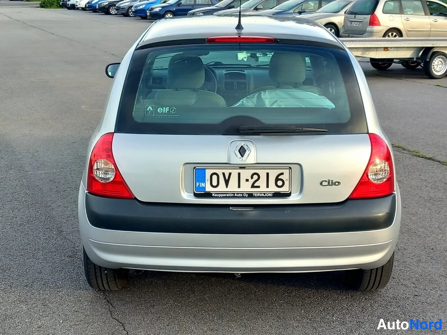Takaa: Koko auton taka- ja sivuosa. Hopeanharmaa Renault Clio -malli takavalolaitteineen ja rekisterikilvenä -216. Kuvassa on nähtävissä avonainen tavaratila jossa var