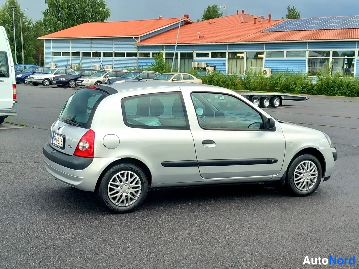 Takaviisto: Hopeanvärinen kaksiovinen piensuvuauto takaviistosta pysäköintialueella, taustalla sininen rakennus ja muita autoja.
