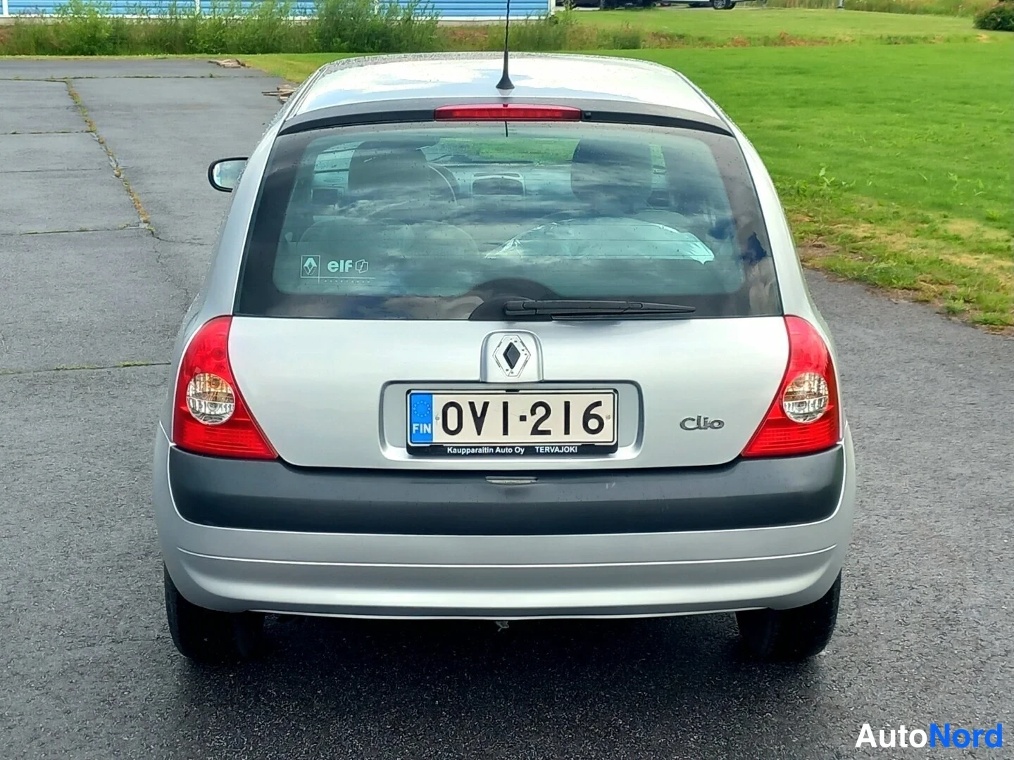 Takaa: Auton takasiluetti: Renault Clio, takavalot ja rekisterikilpi 0VI-216 näkyvät, takalasiin kiinnitetty elf-tarra