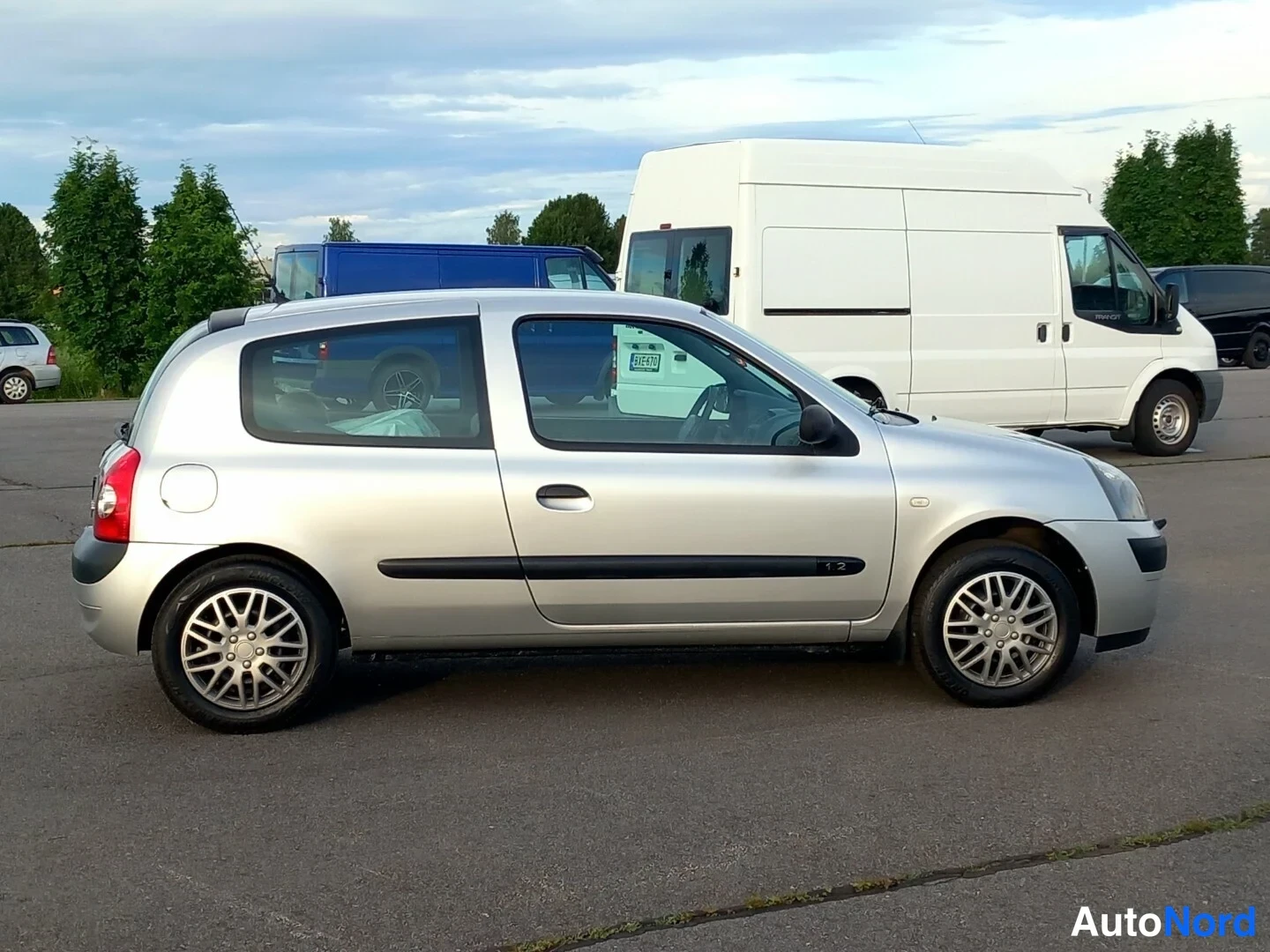 Etuviisto, nokka oikealle: Hopeinen Renault Clio -henkilöauto kolmesta neljäsosasta etuosasta oikealta kuvattuna parkkipaikalla.